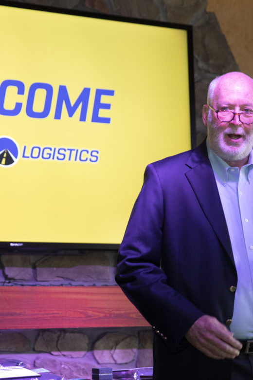 A man stands behind a microphone in front of a screen that says, "Welcome."