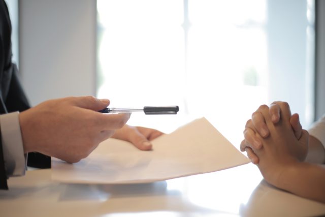 A person holds papers while handing a pen to another person with their hands clasped.