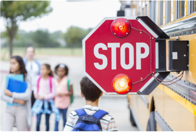 A stop sign swings out from the side of a school bus with children in the background