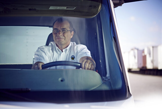 A truck driver sits in the cab of a truck with both hands on the steering wheel