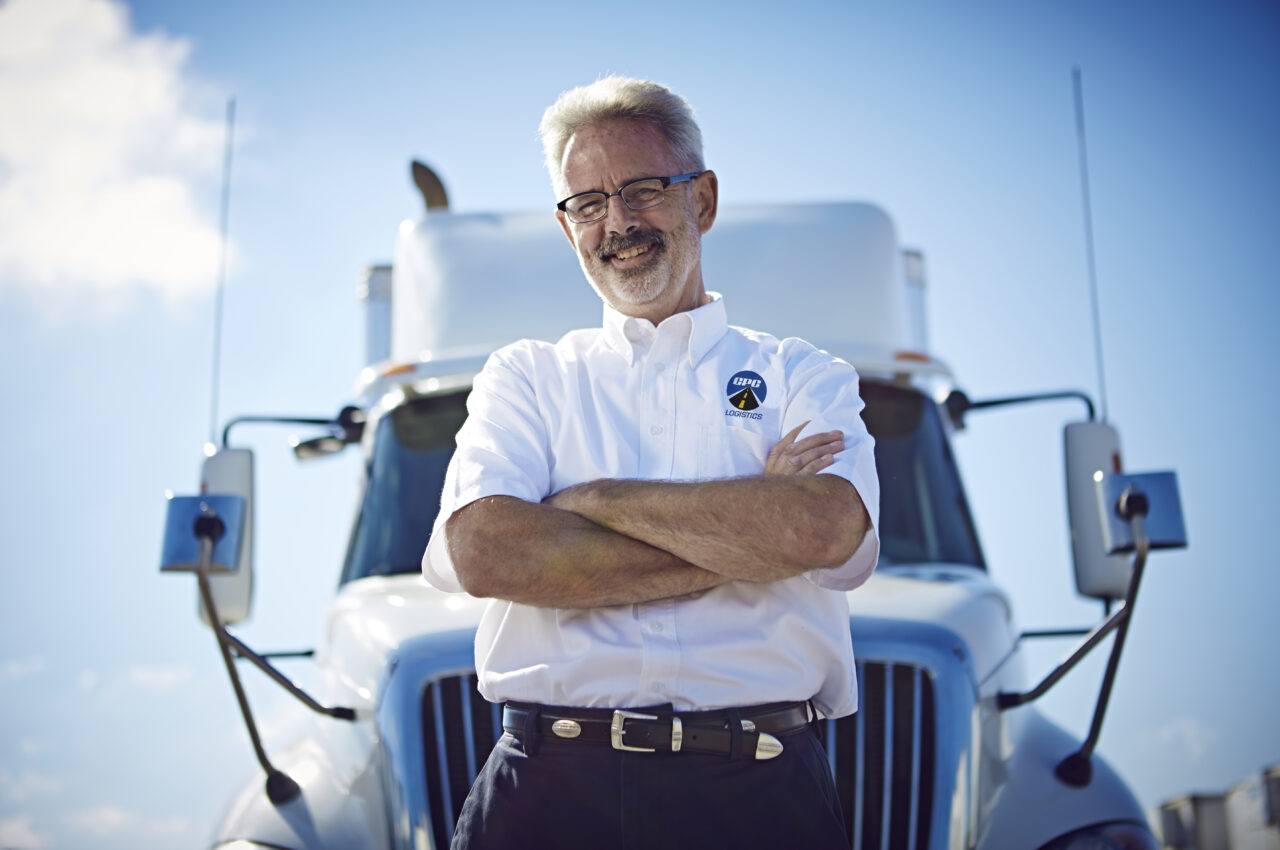 A truck driver stands in front of a semi-truck with his arms crossed.