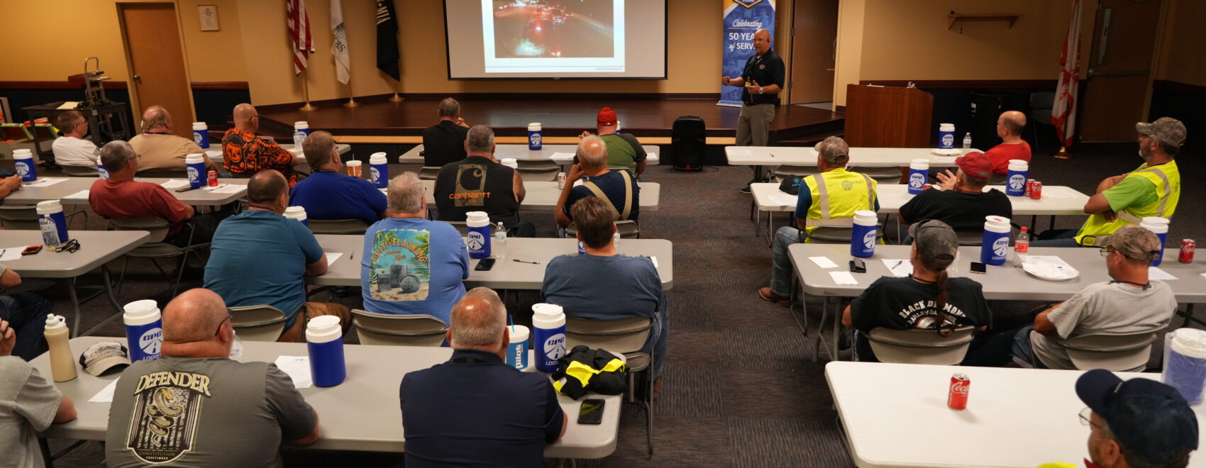 Truck drivers sit in a classroom looking at a man delivering a presentation at the front of the room.