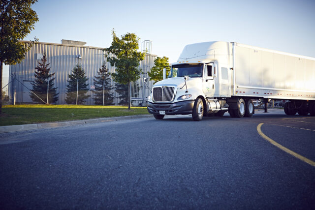 A semi-truck makes a turn on a road in front of a warehouse.