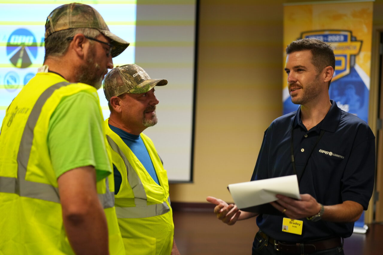 A CPC Logistics manager holding a piece of paper talks with two truck drivers wearing yellow safety vests in a conference room.