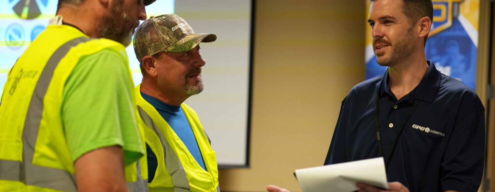 A CPC Logistics manager holding a piece of paper talks with two truck drivers wearing yellow safety vests in a conference room.