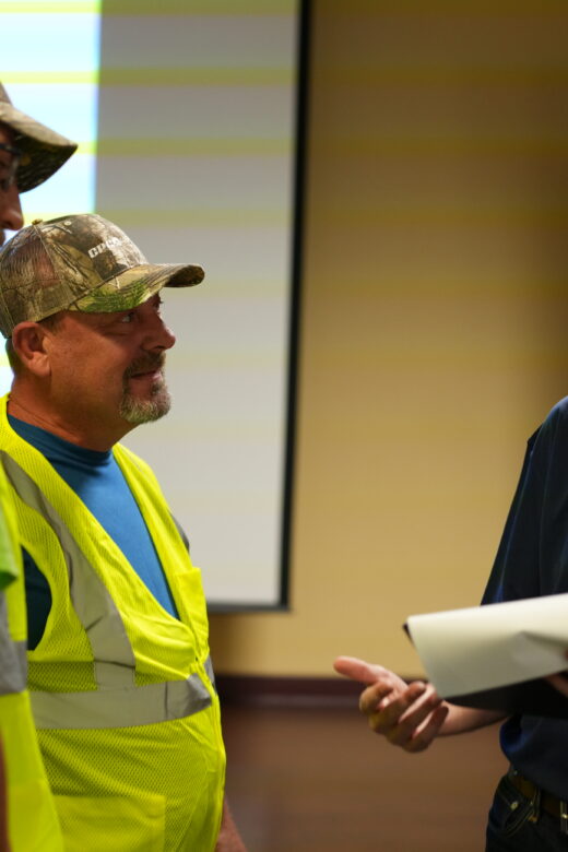 A CPC Logistics manager holding a piece of paper talks with two truck drivers wearing yellow safety vests in a conference room.