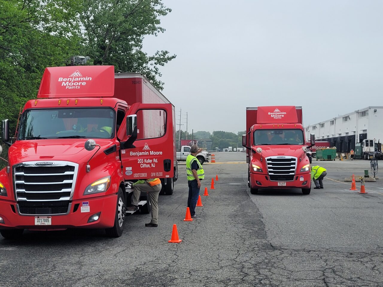 Men in safety vests inspect the exteriors and interiors of three red semi-trucks in a parking lot.
