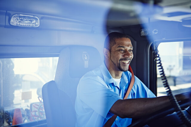A truck driver smiles while sitting behind the wheel of his truck.